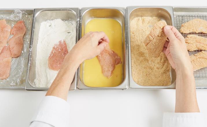 Manos cubriendo piezas de pollo con harina, huevo y pan rallado durante el proceso de preparación de alimentos empanizados.
